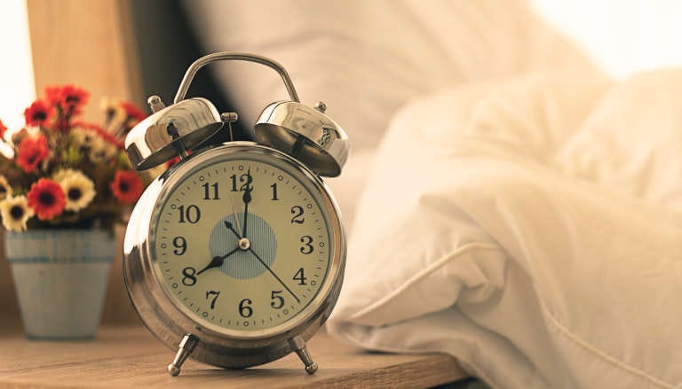 Clock showing 20:00 o'clock, sitting on a bedside table with flowers in the background.