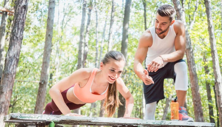 A woman performs push-ups on a bench while a man coaches her in a forested area.