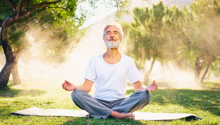 An elderly man meditating outdoors, surrounded by sunlight and trees, radiating a peaceful and serene expression.