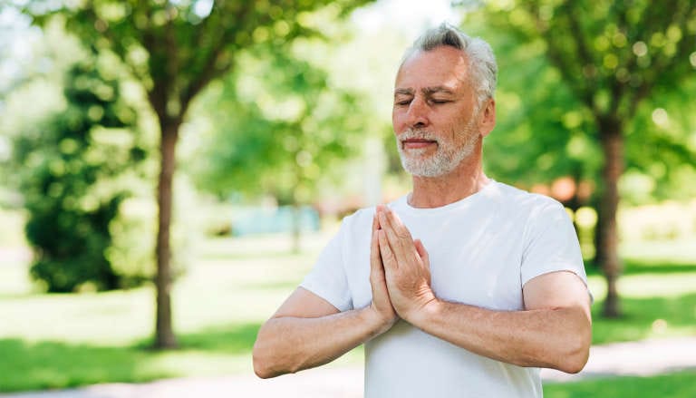 Man meditating outdoors with hands in prayer position, in a green park setting.