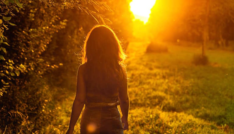 A woman walking along a nature path at golden hour, with sunlight creating a warm, glowing atmosphere through trees and foliage.