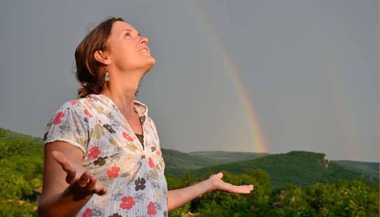 Woman looking up with arms outstretched, a rainbow in the background.