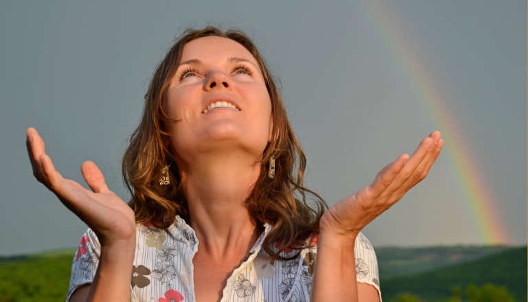 A smiling woman looks upward with open hands, a rainbow visible in the background.