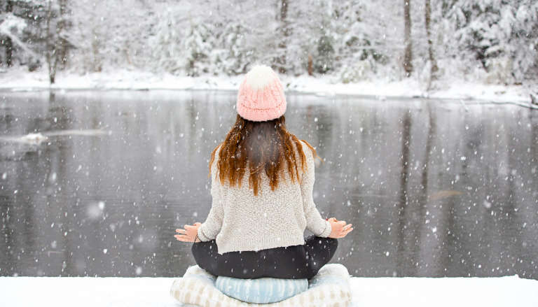 Woman meditating peacefully by a lake in falling snow, embodying the harmony of breath, cold, and mindfulness.