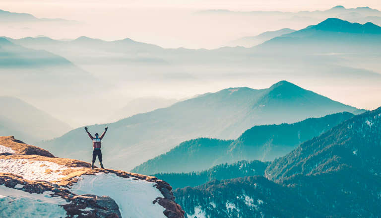 Person with arms raised triumphantly on snowy mountain peak overlooking misty layered mountain ranges at sunrise.