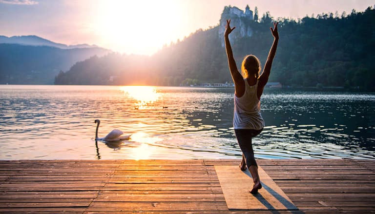 A person practicing yoga on a lakeside dock at sunrise, with a swan gliding in the water.