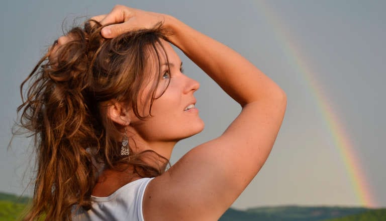 A woman gazing upward with her hand in her hair, a faint rainbow visible in the background.