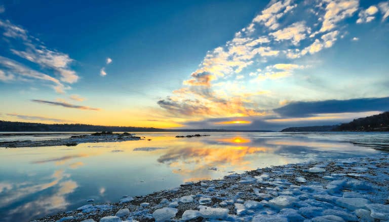 Serene icy lake at golden hour with scattered ice chunks along the shoreline and calm reflective water under warm dawn light.