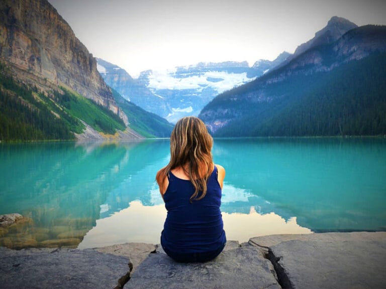 Woman sitting on a stone ledge, back to camera, overlooking a calm turquoise mountain lake at sunset.