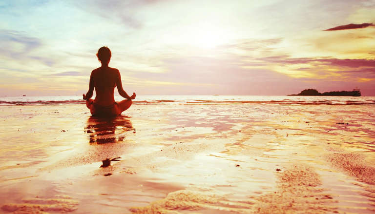 Silhouette of person meditating on beach at sunset, sitting in lotus pose with reflection in wet sand.